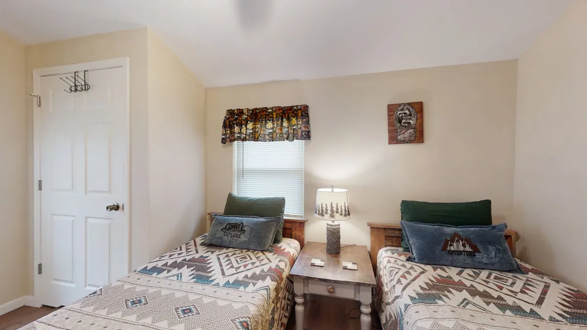 A cheerful twin bedroom featuring two twin beds with patterned quilts, a nightstand with a lamp between them, and nature-inspired wall art. The arrangement provides a friendly, comfortable space ideal for children or singles, supporting group or family stays.