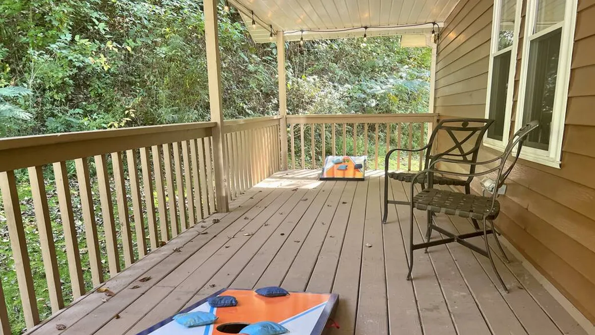 This exterior shot features a wooden deck with cornhole game boards set up and a metal chair, framed by lush greenery. The scene conveys a lively atmosphere perfect for outdoor games, relaxation, and socializing in a natural setting.