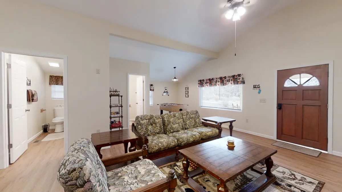 A cozy living area with a plush sofa, coffee table, and a large window framing views of the serene forest outside. The space invites you to enjoy a peaceful morning with coffee or share intimate conversations as natural light spills in. The front door offers a glimpse of the outdoors, suggesting easy access to woodland adventures.