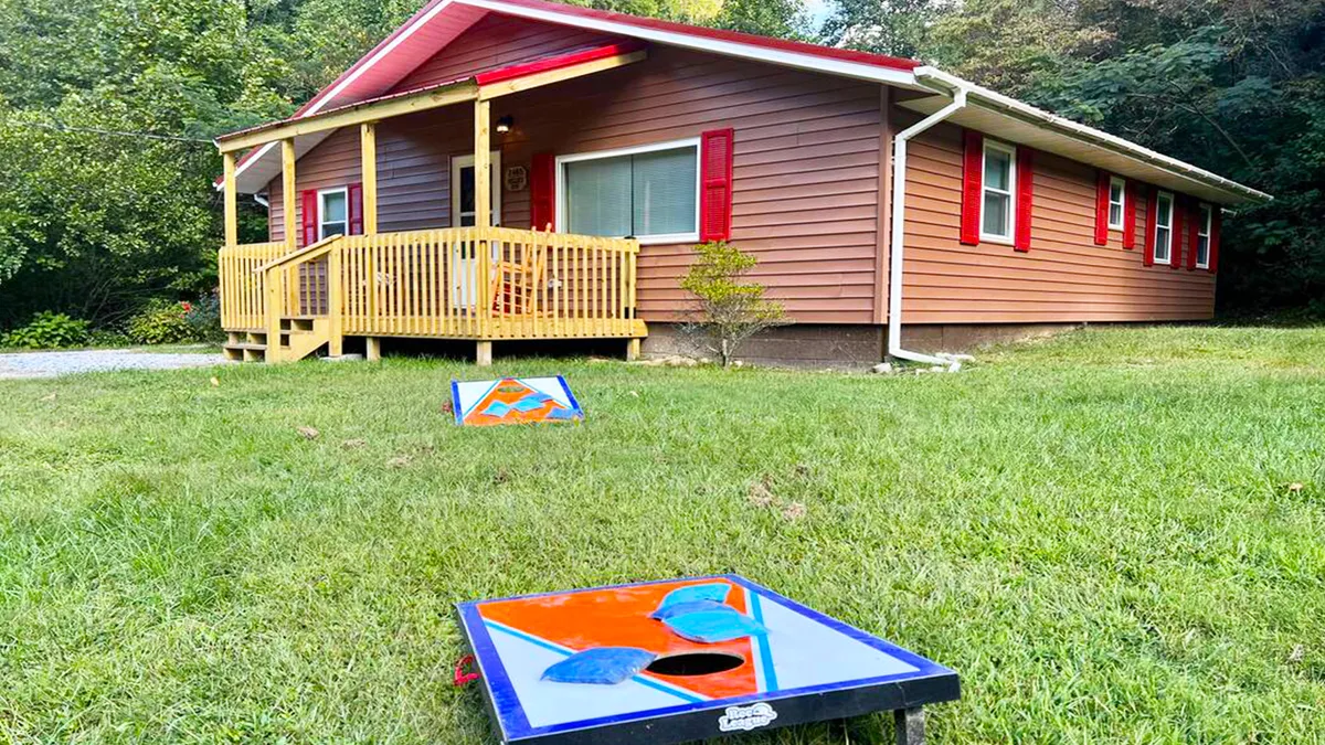 The image showcases a cozy cabin with red shutters nestled among trees, featuring a charming front porch where guests can relax. In the yard, a cornhole setup invites friendly outdoor competitions, with the peaceful ambiance of the Smoky Mountains all around.