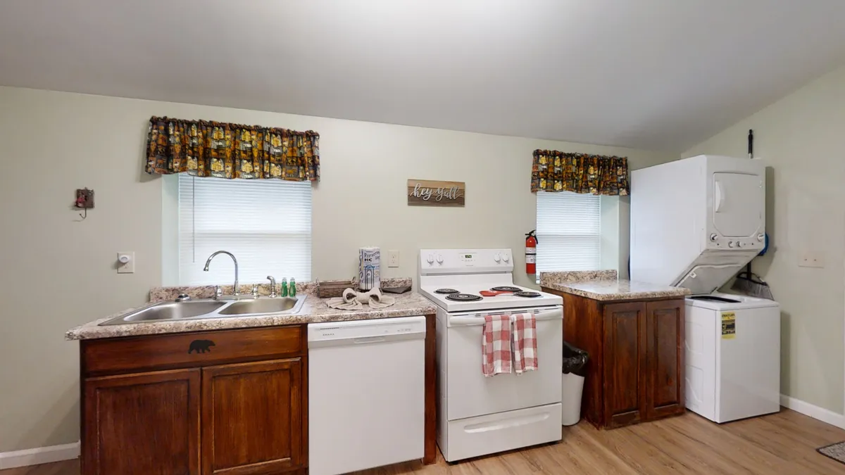 A compact yet highly functional laundry area within the kitchen, featuring a laundry machine, kitchen sink, stove, dishwasher, and window with patterned curtains. The space is both practical and homey, emphasizing ease for families.