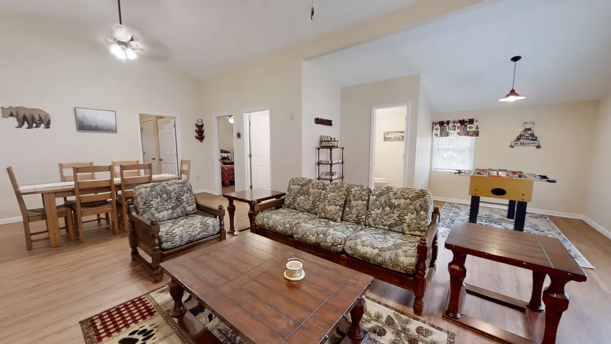 A warm, welcoming living room setup with a floral-patterned sofa, wooden coffee table, a foosball table for games, and a dining table with chairs visible. The open concept encourages group interaction and provides plenty of space for both relaxation and entertainment.