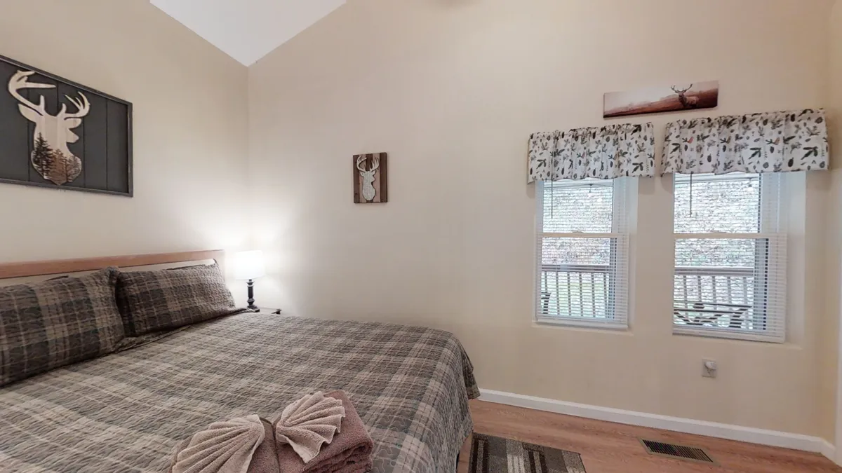 A comfortable queen bed dressed in plaid bedding sits beneath windows adorned with patterned valances, with soft morning light filtering in. Decorative wall art featuring deer adds a playful, romantic touch, while the setting evokes a sense of seclusion and peacefulness—ideal for couples seeking retreat.