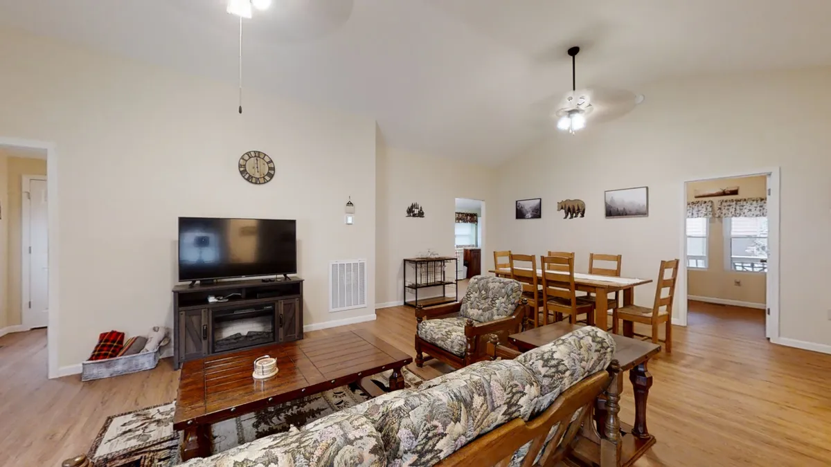 A cozy living room scene featuring a plush floral-upholstered sofa, a dining table in the background, and a large TV unit. The layout suggests an open flow between the living, dining, and entertainment areas, capturing the central hub where guests can relax, watch movies, or gather for meals.