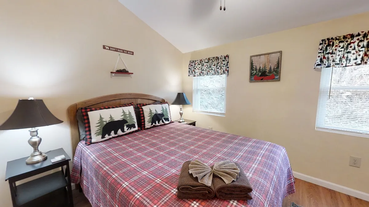 A warmly decorated bedroom features a bed with patterned blankets and pillows depicting bears and trees, a bedside lamp, and a wall art of a forest scene. The setup feels inviting, perfect for snuggling up after a day exploring nearby attractions. Natural light and rustic decor create a cozy hideaway vibe.