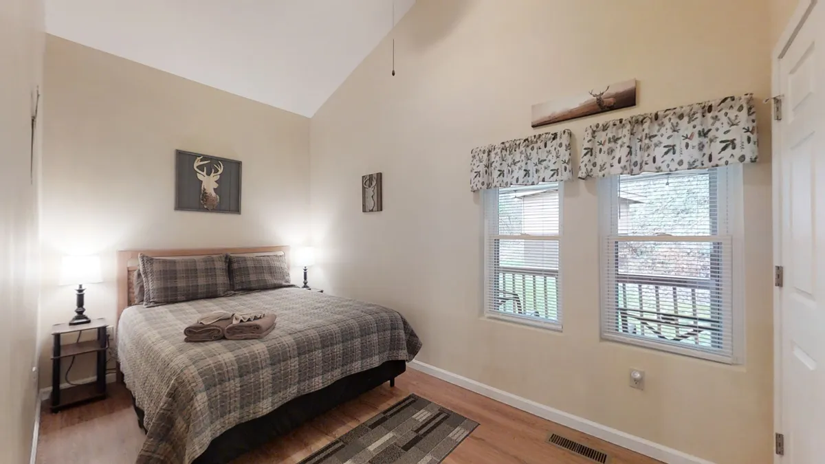 A welcoming bedroom with a bed dressed in plaid bedding, decorative wall art featuring a deer, two bedside lamps, and windows with decorative curtains. The soft lighting and tranquil atmosphere evoke a sense of restful comfort and privacy, ideal for couples or solo travelers.
