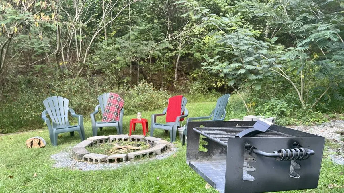 This image captures a cozy fire pit area surrounded by adirondack chairs with a charcoal grill nearby, all nestled within lush greenery. The setup invites gatherings for campfire storytelling, grilling, and relaxing outdoors, highlighting the space as a social hub for both day and evening enjoyment.